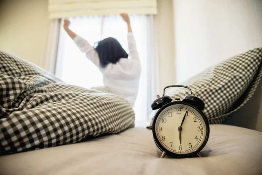 A woman just woke up with stretched arms and an alarm clock at 6:04 a.m. in the concept of waking early for the moving day