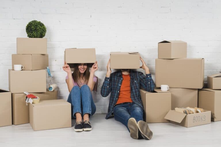 A young couple hiding their faces with moving carton in the concept of 'What Are the Hidden Costs in Home Removals Services'.
